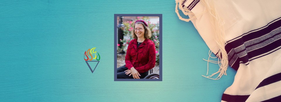 Julia Watts Belser, a white Jewish woman with curly brown hair, sits happily in her wheelchair in front of a pink flowering bush. She's wearing a patterned red blazer and red kippah (beret) to match.